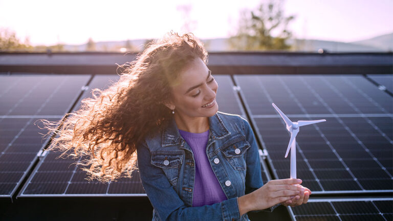 Mulher na frente de uma campo de painéis solares, segurando uma miniatura de pá eólica