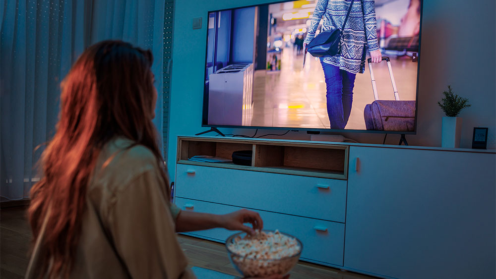 Mulher assistindo TV em sala moderna com pipoca, representando conforto e imersão de um home theater em casa.